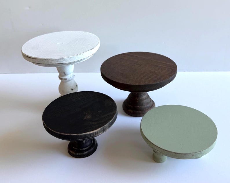 Puede incluir: Cuatro soportes circulares de madera con pedestal de diferentes alturas y colores. Un soporte es blanco con acabado desgastado, uno es marr&oacute;n oscuro, uno es negro y otro es de color verde salvia suave. Cada soporte tiene una base redonda.