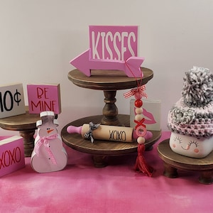 May include: A tiered wooden stand with pink and white Valentine's Day decorations. The top tier has a pink arrow with the word "KISSES" on it. The middle tier has a rolling pin with the word "XOXO" on it. The bottom tier has a snowman with a pink ribbon and a pink and white sign that says "BE MINE".