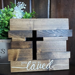 May include: Wooden sign with a cross cutout and the text "you are so loved" in white script. The sign is made of dark brown wood planks.