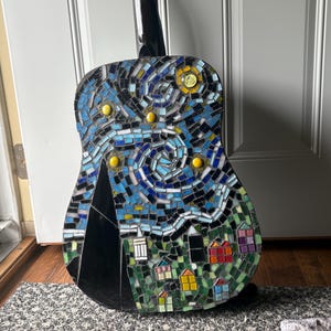 K&ouml;nnte beinhalten: Akustikgitarre mit einem Mosaikdesign, das eine Sternennachtszene darstellt. Das Mosaik zeigt blaue, gelbe und schwarze Fliesen mit einer Dorfszene am unteren Rand. Der Hals der Gitarre ist schwarz.