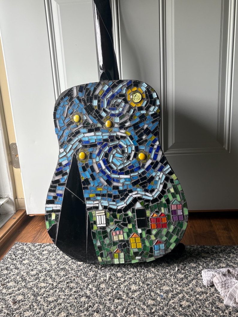K&ouml;nnte beinhalten: Akustikgitarre mit einem Mosaikdesign, das eine Sternennachtszene darstellt. Das Mosaik zeigt blaue, gelbe und schwarze Fliesen mit einer Dorfszene am unteren Rand. Der Hals der Gitarre ist schwarz.
