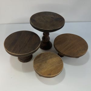 Puede incluir: Un conjunto de cuatro soportes circulares de madera con un acabado te&ntilde;ido en marr&oacute;n oscuro. La colecci&oacute;n incluye un soporte de pedestal alto con base torneada y tres soportes m&aacute;s cortos de superficie plana y alturas variadas.
