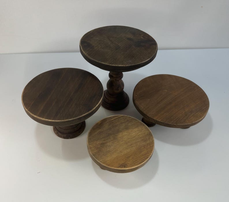 Puede incluir: Un conjunto de cuatro soportes circulares de madera con un acabado te&ntilde;ido en marr&oacute;n oscuro. La colecci&oacute;n incluye un soporte de pedestal alto con base torneada y tres soportes m&aacute;s cortos de superficie plana y alturas variadas.