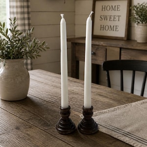 Peut inclure: Deux bougies blanches effil&eacute;es dans des bougeoirs en bois brun fonc&eacute; sont pos&eacute;es sur une table en bois rustique. Un vase avec de la verdure et une enseigne encadr&eacute;e avec l'inscription &laquo; HOME SWEET HOME &raquo; sont en arri&egrave;re-plan.