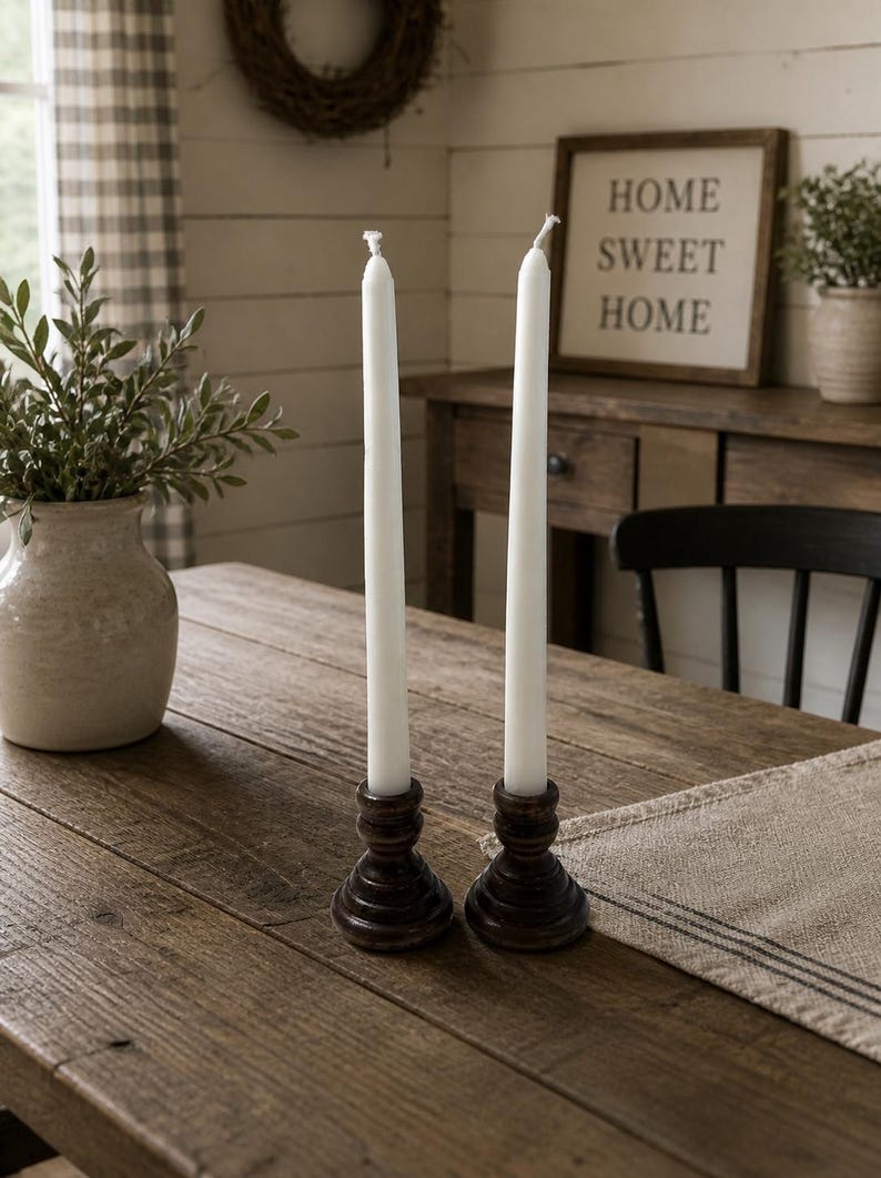 Peut inclure: Deux bougies blanches effil&eacute;es dans des bougeoirs en bois brun fonc&eacute; sont pos&eacute;es sur une table en bois rustique. Un vase avec de la verdure et une enseigne encadr&eacute;e avec l'inscription &laquo; HOME SWEET HOME &raquo; sont en arri&egrave;re-plan.