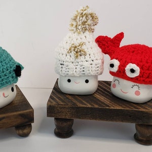 May include: Three white ceramic mugs with different crocheted hats. The mugs have a happy face and are sitting on wooden stands. The hats are a red crab, a white beanie with a gold pom-pom, and a teal narwhal.