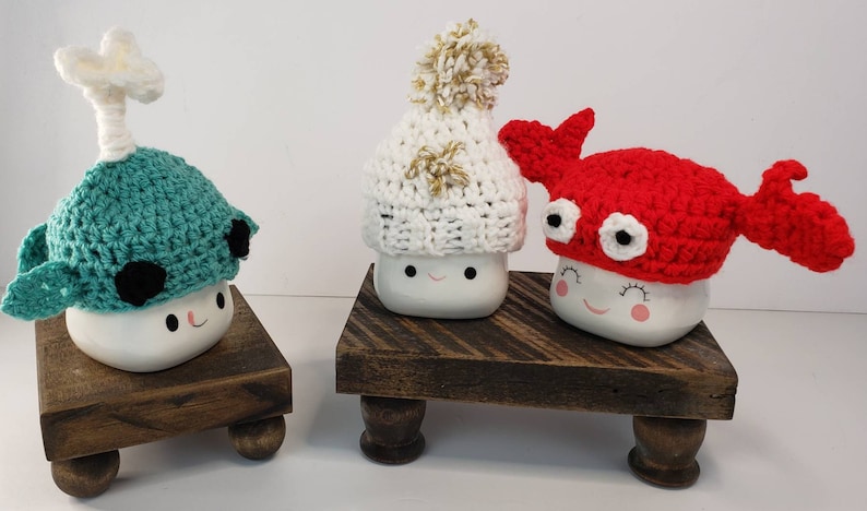 May include: Three white ceramic mugs with different crocheted hats. The mugs have a happy face and are sitting on wooden stands. The hats are a red crab, a white beanie with a gold pom-pom, and a teal narwhal.