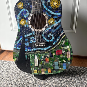 K&ouml;nnte beinhalten: Akustikgitarre mit einem Mosaikdesign, inspiriert von Van Goghs "Sternennacht". Die Gitarre hat einen dunkelblauen Hintergrund mit wirbelnden blauen, gelben und wei&szlig;en Mosaikfliesen. Der untere Teil zeigt eine Dorfszene mit bunten Geb&auml;uden.