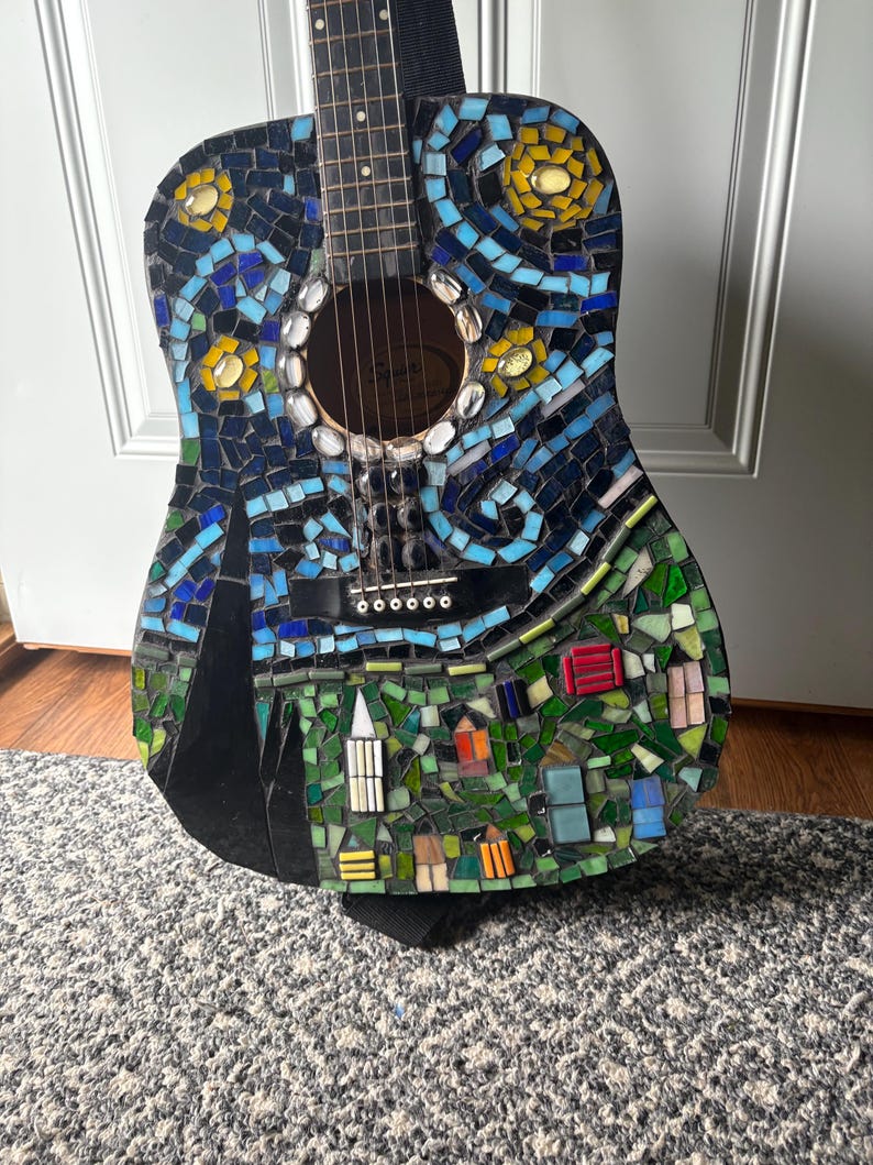 K&ouml;nnte beinhalten: Akustikgitarre mit einem Mosaikdesign, inspiriert von Van Goghs "Sternennacht". Die Gitarre hat einen dunkelblauen Hintergrund mit wirbelnden blauen, gelben und wei&szlig;en Mosaikfliesen. Der untere Teil zeigt eine Dorfszene mit bunten Geb&auml;uden.