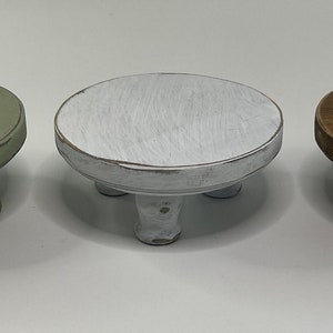 Puede incluir: Tres soportes redondos de madera peque&ntilde;os con patas cortas torneadas. Un soporte est&aacute; pintado de verde salvia, otro de blanco con un acabado desgastado y el tercero tiene un tinte de madera marr&oacute;n oscuro natural. Cada base tiene una superficie plana.