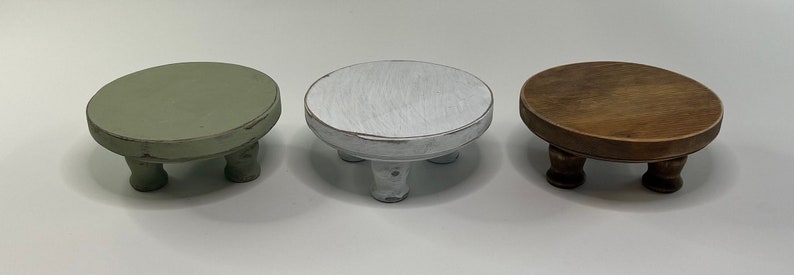 Puede incluir: Tres soportes redondos de madera peque&ntilde;os con patas cortas torneadas. Un soporte est&aacute; pintado de verde salvia, otro de blanco con un acabado desgastado y el tercero tiene un tinte de madera marr&oacute;n oscuro natural. Cada base tiene una superficie plana.