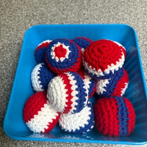 May include: A collection of red, white, and blue crocheted balls in a blue plastic container. The balls are in various patterns, some with stripes and others with a star-like design in the center. The balls are likely handmade.