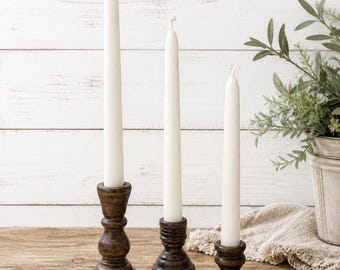 May include: Three white taper candles in varying heights, set in dark brown wooden candle holders. The candle holders have a detailed, turned design. The candles are on a wooden surface with a white wood-paneled background.