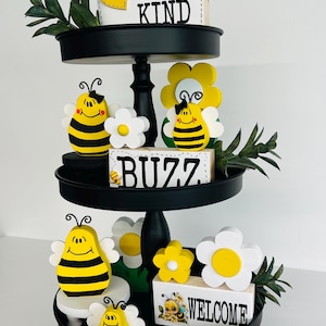 May include: A three-tiered black tray with bee and flower decorations. The top tier has a sign that says "BEE KIND". The middle tier has a sign that says "BUZZ". The bottom tier has a sign that says "WELCOME". The decorations are yellow, white, and black.