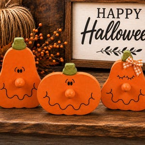 May include: Three orange wooden pumpkin decorations with green stems and painted black facial features. Each pumpkin has a round wooden nose. The right pumpkin features a small orange and white checkered bow attached to its green stem.