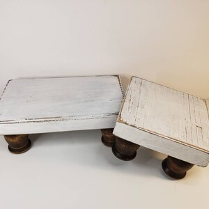 May include: Two white wooden risers with distressed paint and turned wooden legs. The risers are different sizes and are stacked on top of each other.