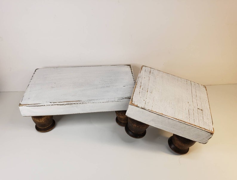 May include: Two white wooden risers with distressed paint and turned wooden legs. The risers are different sizes and are stacked on top of each other.