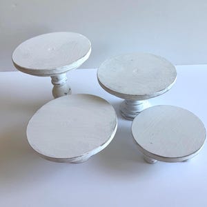 Puede incluir: Un conjunto de cuatro elevadores de madera blanca con un acabado desgastado. Dos elevadores cuentan con bases de pedestal altas y torneadas, mientras que los otros dos son m&aacute;s cortos con pies bajos y redondeados. Cada uno tiene una base circular.