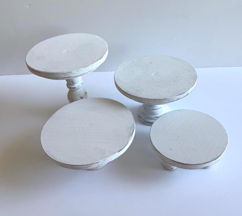 Puede incluir: Un conjunto de cuatro elevadores de madera blanca con un acabado desgastado. Dos elevadores cuentan con bases de pedestal altas y torneadas, mientras que los otros dos son m&aacute;s cortos con pies bajos y redondeados. Cada uno tiene una base circular.