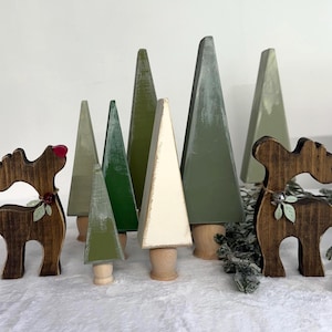 May include: A collection of wooden Christmas decorations, including stylized trees in various shades of green and a cream color, and two reindeer. The reindeer are brown with red accents and floral details. The trees are various heights.