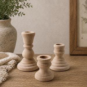 Peut inclure: Trois bougeoirs en bois non finis de hauteurs variables sont pos&eacute;s sur une surface en bois. Les bougeoirs ont un design tourn&eacute; avec une base circulaire et un dessus encastr&eacute; pour les bougies. Un vase avec de la verdure est en arri&egrave;re-plan.
