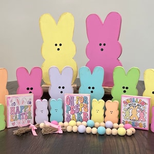 May include: A collection of pastel-coloured bunny-shaped wooden figurines with black eyes and mouths. The figurines are arranged in a row with signs that say "Happy Easter", "Oh For Peps Sake", and "Chillin With My Peeps".
