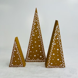 May include: Three decorative, cone-shaped Christmas trees. The trees are painted a warm brown color and decorated with white icing-like zigzags and dots. The trees vary in size, creating a layered effect.