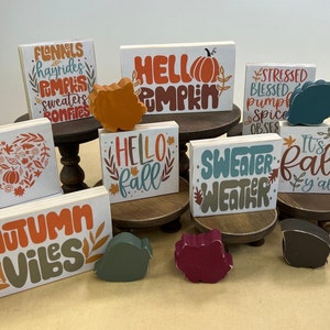 May include: A collection of eight wooden signs with autumn-themed sayings, including "Hello Pumpkin", "Sweater Weather", "It's Fall Y'all", and "Autumn Vibes". The signs are decorated with fall colors and leaves.