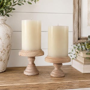 May include: Two ivory-colored pillar candles sit on light brown wooden candle holders. The candle holders have a pedestal base and a round top. The candles are unscented and cylindrical, creating a warm, inviting ambiance.