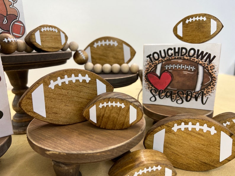 May include: Wooden tiered stand decorated with footballs and a sign that says "Touchdown Season". The sign has a red heart and a brown football on a leopard print background.