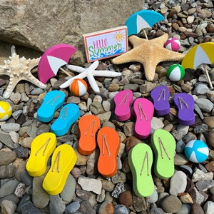 May include: A collection of colorful flip flops, beach balls, starfish, and umbrellas on a bed of gray and brown pebbles. The sign reads "Hello Summer".