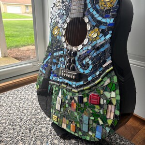 K&ouml;nnte beinhalten: Akustikgitarre mit einem Mosaikdesign, inspiriert von Van Goghs "Sternennacht". Das Mosaik zeigt blaue, gelbe und gr&uuml;ne Fliesen, die einen Nachthimmel und eine Dorfszene darstellen. Die Gitarre hat einen schwarzen Korpus und einen schwarzen Gurt.