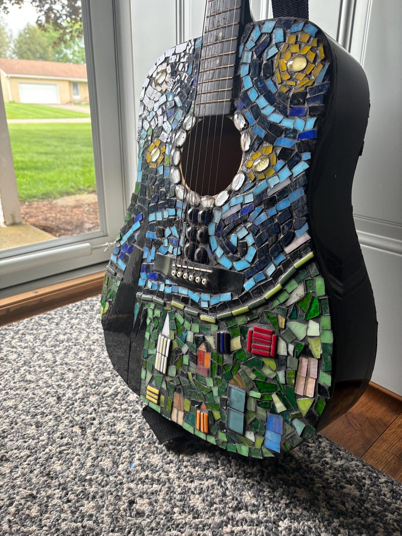 K&ouml;nnte beinhalten: Akustikgitarre mit einem Mosaikdesign, inspiriert von Van Goghs "Sternennacht". Das Mosaik zeigt blaue, gelbe und gr&uuml;ne Fliesen, die einen Nachthimmel und eine Dorfszene darstellen. Die Gitarre hat einen schwarzen Korpus und einen schwarzen Gurt.