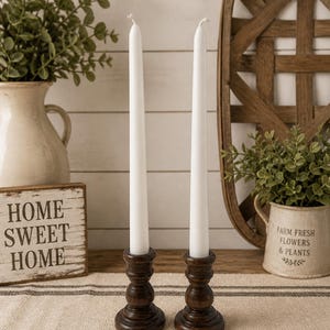 Peut inclure: Deux bougies blanches effil&eacute;es dans des bougeoirs en bois brun fonc&eacute;. Les bougeoirs ont un design tourn&eacute; d&eacute;coratif. Les bougies sont plac&eacute;es sur un chemin de table en tissu ray&eacute;. Un panneau en bois avec le texte "HOME SWEET HOME" est visible.