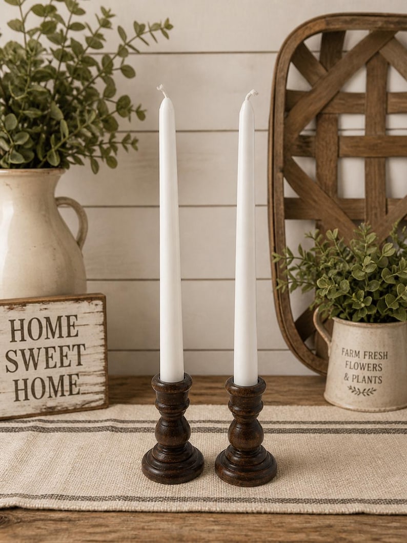 Peut inclure: Deux bougies blanches effil&eacute;es dans des bougeoirs en bois brun fonc&eacute;. Les bougeoirs ont un design tourn&eacute; d&eacute;coratif. Les bougies sont plac&eacute;es sur un chemin de table en tissu ray&eacute;. Un panneau en bois avec le texte "HOME SWEET HOME" est visible.