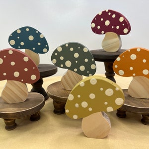 May include: Six wooden mushroom figurines with white polka dots painted on them. Each mushroom is on a small wooden pedestal with three legs. The mushrooms are painted in different colors: red, teal, green, orange, yellow, and burgundy.