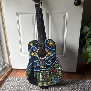 K&ouml;nnte beinhalten: Akustikgitarre mit Mosaikdesign. Der Korpus weist ein farbenfrohes Mosaikmuster mit blauen, gelben, gr&uuml;nen und schwarzen Fliesen auf, das eine Landschaft mit Geb&auml;uden und Blumen darstellt. Die Gitarre hat einen schwarzen Hals und einen schwarzen Gurt.