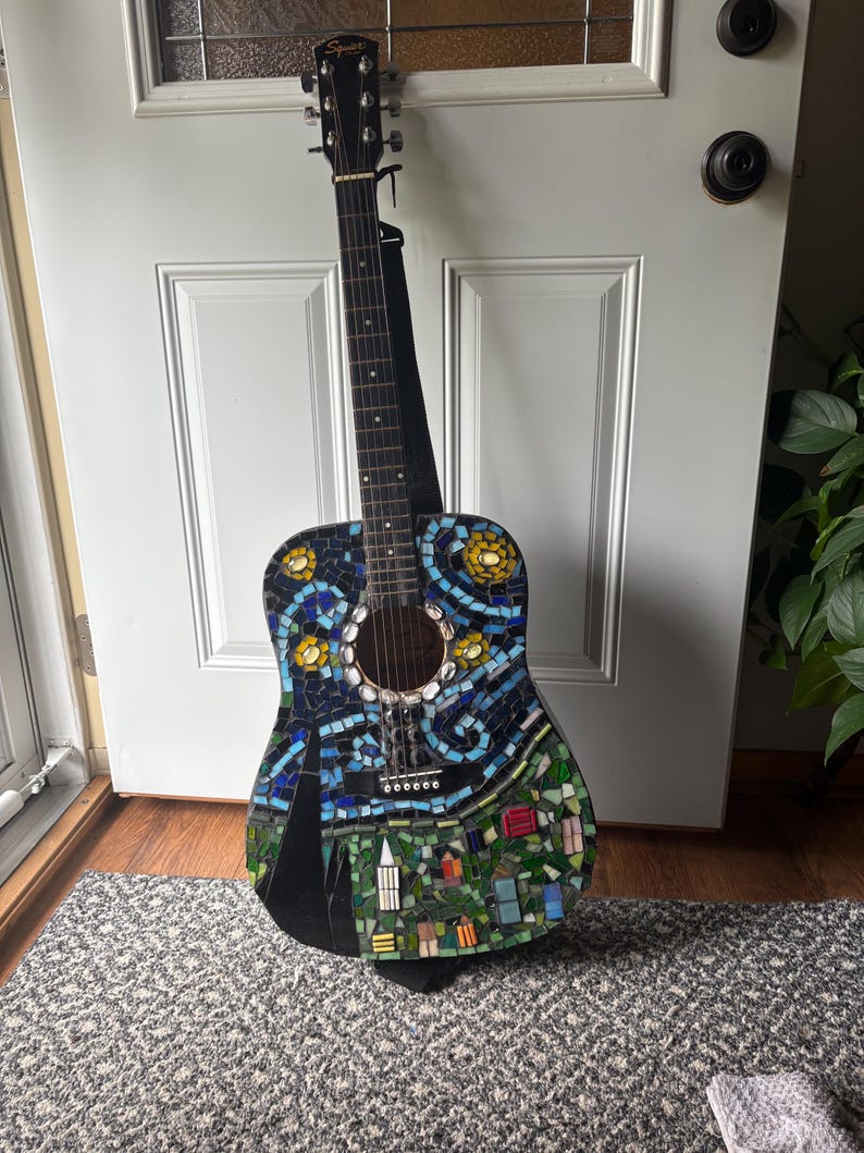 K&ouml;nnte beinhalten: Akustikgitarre mit Mosaikdesign. Der Korpus weist ein farbenfrohes Mosaikmuster mit blauen, gelben, gr&uuml;nen und schwarzen Fliesen auf, das eine Landschaft mit Geb&auml;uden und Blumen darstellt. Die Gitarre hat einen schwarzen Hals und einen schwarzen Gurt.