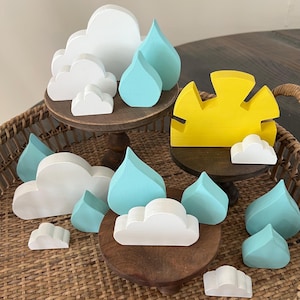 May include: Wooden craft decorations for a nursery or child's room. The decorations include a yellow sun, white clouds, and blue raindrops. The decorations are arranged on tiered wooden stands.