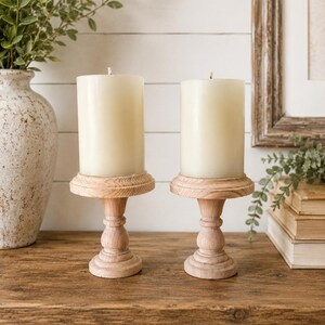 May include: Two ivory-colored pillar candles sit atop matching wooden candle holders. The candle holders have a turned design and are placed on a rustic wooden surface. A decorative vase with greenery is visible in the background.