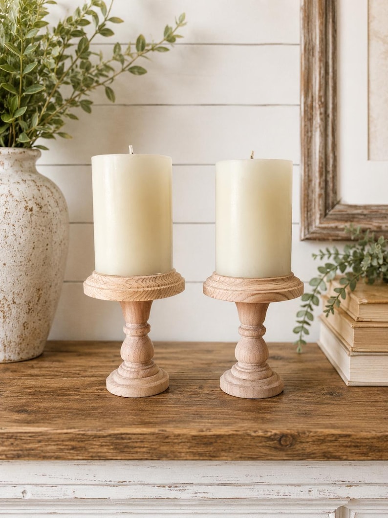 May include: Two ivory-colored pillar candles sit atop matching wooden candle holders. The candle holders have a turned design and are placed on a rustic wooden surface. A decorative vase with greenery is visible in the background.