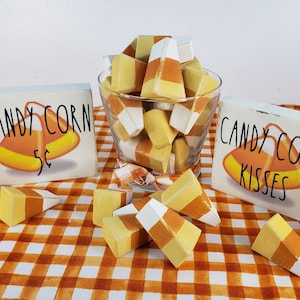 May include: Two wooden signs with the text "Candy Corn 5c" and "Candy Corn Kisses" with a bowl of yellow, white, and orange candy corn in the center. The signs and candy corn are on a checkered orange and white tablecloth.