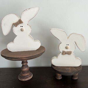 May include: Two white wooden bunny figurines with brown bows, sitting on brown wooden stands. The larger bunny is on a pedestal stand, while the smaller bunny is on a stand with three legs.