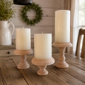 May include: Three cream-colored pillar candles on wooden candle holders. The candle holders have a light wood finish and a pedestal base. The candles are of varying heights, creating a layered display on a wooden table.