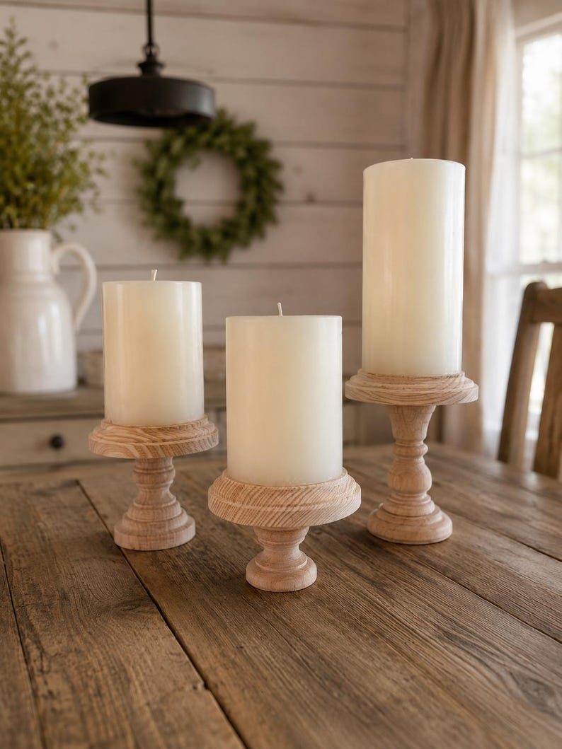 May include: Three cream-colored pillar candles on wooden candle holders. The candle holders have a light wood finish and a pedestal base. The candles are of varying heights, creating a layered display on a wooden table.