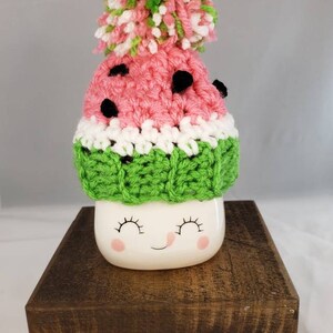 May include: A white ceramic mug with a smiling face and pink cheeks, wearing a crocheted watermelon hat with a green and white pom-pom. The mug is sitting on a wooden stand with rounded legs.