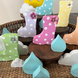 May include: A set of wooden rain boot figurines with polka dots and flowers. The boots are in various colors, including pink, blue, green, yellow, and purple. The set also includes wooden clouds and raindrops.