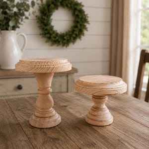 May include: Two unfinished wooden pedestal stands with a natural wood grain finish. One stand is taller with a detailed top edge, while the other is shorter. Both are on a wooden table, suggesting a rustic or farmhouse style.