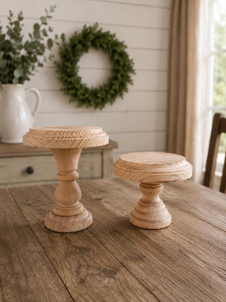 May include: Two unfinished wooden pedestal stands with a natural wood grain finish. One stand is taller with a detailed top edge, while the other is shorter. Both are on a wooden table, suggesting a rustic or farmhouse style.
