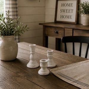 Peut inclure: Une table en bois rustique pr&eacute;sente trois bougeoirs blancs patin&eacute;s. Un vase avec de la verdure et une enseigne encadr&eacute;e avec l'inscription "HOME SWEET HOME" sont &eacute;galement visibles. L'ensemble &eacute;voque une ambiance de ferme chaleureuse.